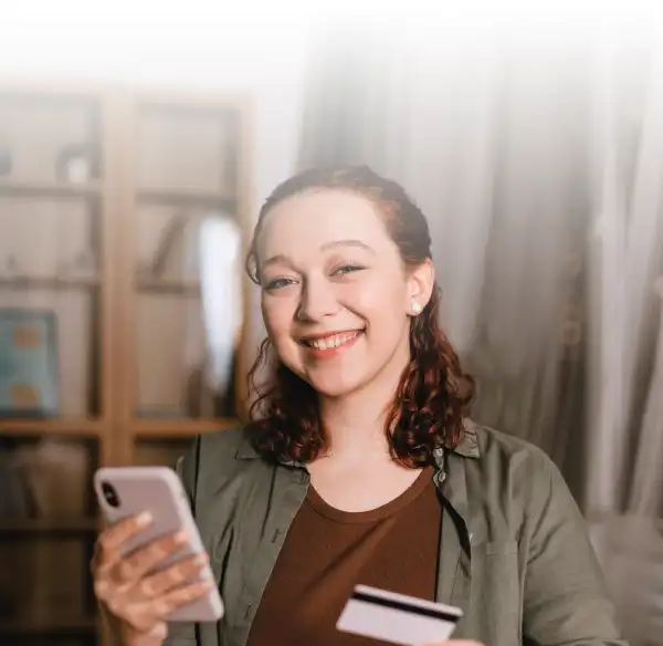 chica jóven sonriendo con un celular en una mano y una tarjeta de crédito en la otra simulando un pago online